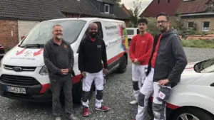Vier Männer in Arbeitskleidung und Jacken stehen draußen vor zwei Arbeitswagen. Hinter ihnen stehen Häuser mit Ziegeldächern und Kies auf dem Boden. Die Lieferwagen sind mit Firmenlogos und Texten beschriftet.