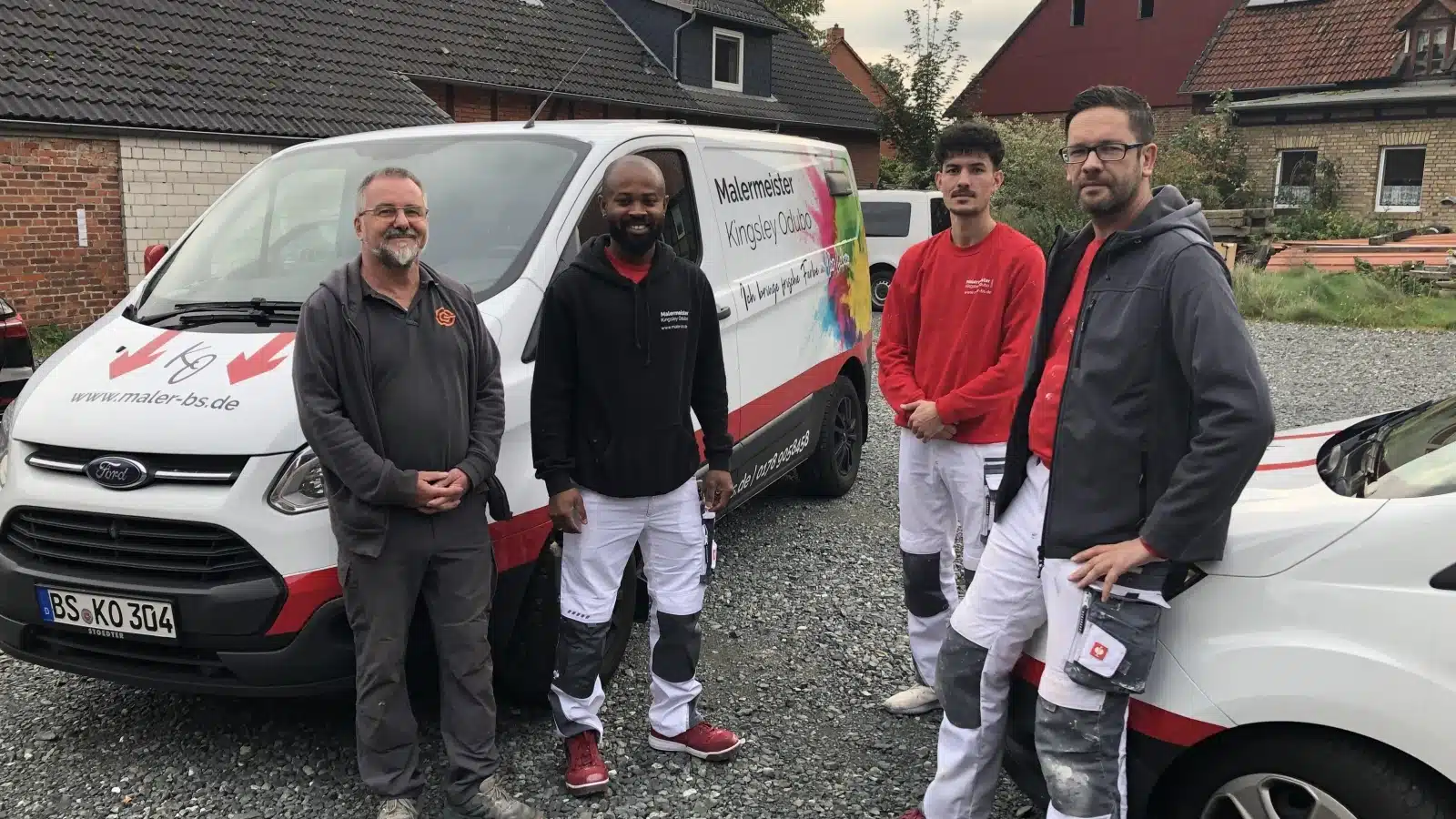 Vier Männer in Arbeitskleidung und Jacken stehen draußen vor zwei Arbeitswagen. Hinter ihnen stehen Häuser mit Ziegeldächern und Kies auf dem Boden. Die Lieferwagen sind mit Firmenlogos und Texten beschriftet.